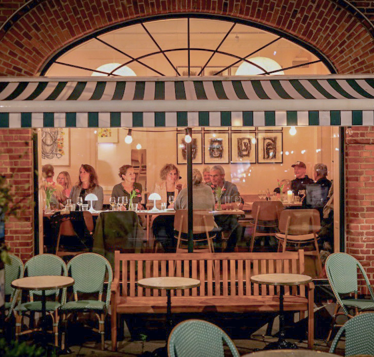 Restaurant set udefra om aftenen gennem stort vindueparti. Der er lys og mennesker inde i restauranten.